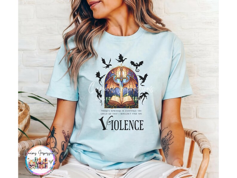 May include: Light blue t-shirt featuring a fantasy-themed graphic. The design includes a sword, open book, stained glass window, and dragons. The word "VIOLENCE" is printed below the graphic. The shirt has short sleeves.