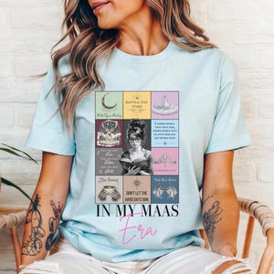 K&ouml;nnte beinhalten: Hellblaues T-Shirt mit einer farbenfrohen Grafik, die eine Frau beim Lesen eines Buches zeigt, umgeben von Illustrationen und Text zum Thema B&uuml;cher. Der Text "IN MY MAAS Era" ist in einer stilvollen Schriftart gedruckt.