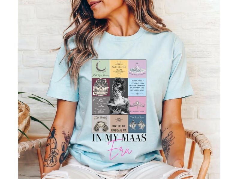 K&ouml;nnte beinhalten: Hellblaues T-Shirt mit einer farbenfrohen Grafik, die eine Frau beim Lesen eines Buches zeigt, umgeben von Illustrationen und Text zum Thema B&uuml;cher. Der Text "IN MY MAAS Era" ist in einer stilvollen Schriftart gedruckt.