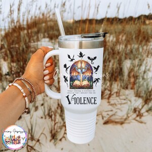 May include: White travel mug with a handle, featuring a design with a sword, open book, and bats. The word "Violence" is printed below the design. Includes a straw and a black lid.