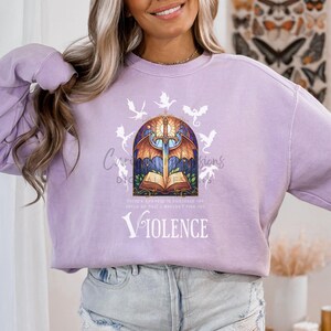 May include: A light purple sweatshirt featuring a graphic design with a sword, open book, and dragon illustrations. The word "VIOLENCE" is printed in white. The design is centered on the front of the sweatshirt.