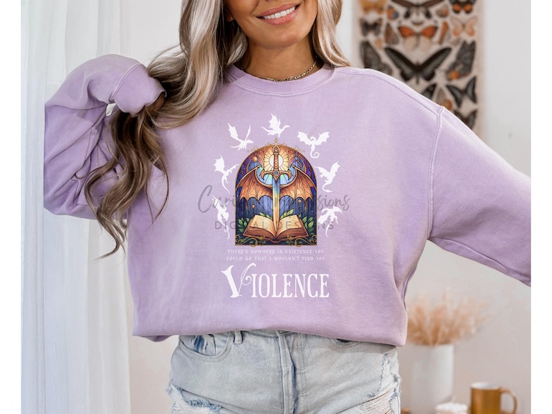 May include: A light purple sweatshirt featuring a graphic design with a sword, open book, and dragon illustrations. The word "VIOLENCE" is printed in white. The design is centered on the front of the sweatshirt.