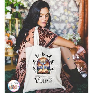 May include: A white tote bag featuring a fantasy-themed design. The design includes a sword, open book, and dragon silhouettes. The word "VIOLENCE" is printed below the illustration. The bag is held by a person.