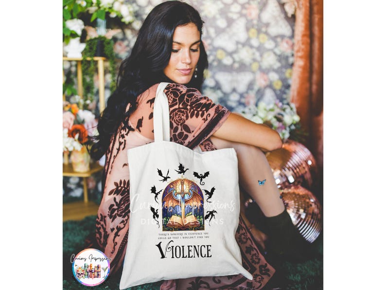 May include: A white tote bag featuring a fantasy-themed design. The design includes a sword, open book, and dragon silhouettes. The word "VIOLENCE" is printed below the illustration. The bag is held by a person.