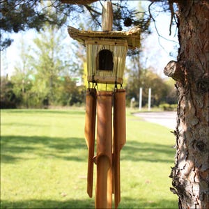 May include: A bamboo wind chime with a small, house-shaped top and several long, cylindrical chimes. The top has a thatched roof and a small arched opening. The chimes are suspended from the top with black string. The background is a green lawn and trees.