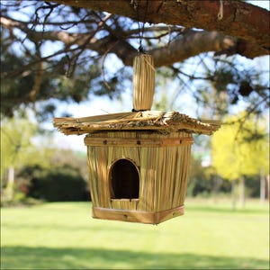 May include: A rustic birdhouse, crafted from natural reeds and wood, suspended from a tree branch. The birdhouse has a square body with a circular entrance and a thatched roof. The structure is a light brown colour, blending with the natural surroundings.