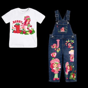 May include: A white t-shirt and denim dungarees set with a Strawberry Shortcake theme. The t-shirt has the text "Berry 1st" with a Strawberry Shortcake graphic. The dungarees have Strawberry Shortcake graphics, the letter "J" and the number "1".