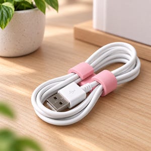Op de afbeelding: Een witte USB-oplaadkabel opgerold in een ovale vorm en vastgezet met twee lichtroze siliconen kabelbinders. De kabel is voorzien van een standaard USB-A-connector met een metallic zilveren punt en een witte kunststof behuizing met een USB-symbool.