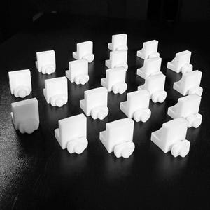 Op de afbeelding: Een verzameling kleine, witte, 3D-geprinte objecten. Elk object heeft een rechthoekige bovenkant en een afgeronde basis, die lijken op miniatuur architecturale elementen of decoratieve stukken. De objecten zijn op een donker oppervlak geplaatst.