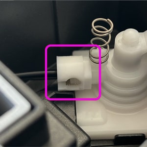 Peut inclure: Gros plan sur une pièce en plastique blanche avec un ressort attaché. La pièce a un petit trou au centre et est probablement un composant d'un dispositif mécanique.