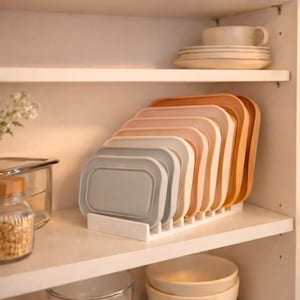 Peut inclure: Un ensemble de couvercles rectangulaires pour récipients alimentaires, dans des tons neutres comme le gris, le beige et le pêche, est rangé dans un support blanc. Les couvercles sont disposés dans un placard blanc, pour l'organisation de la cuisine.