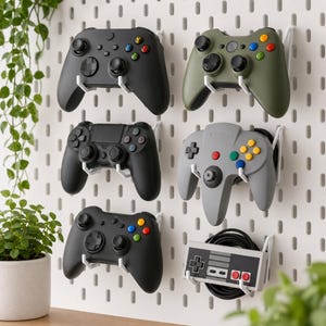 Op de afbeelding: Een wit geperforeerd wandbord als houder voor vijf videogamecontrollers en één rechthoekige NES-controller met een opgerold zwart snoer. De controllers zijn zwart, olijfgroen en grijs en hebben diverse knoppen, joysticks en richtingsknoppen.