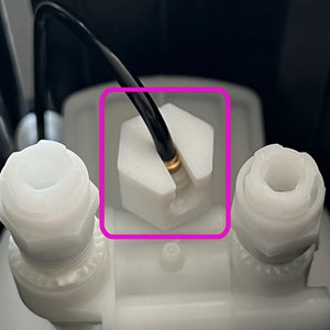 Könnte beinhalten: Nahaufnahme eines weißen Kunststoff-Connectors mit einem schwarzen Kabel. Der Connector hat eine sechseckige Form und ein goldfarbenes Metallstück in der Mitte.