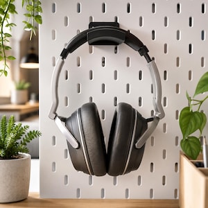 Könnte beinhalten: Over-Ear-Kopfhörer mit schwarzen gepolsterten Ohrmuscheln und einem silbernen Metallrahmen, aufgehängt an einem schwarzen Kunststoffhaken an einer weißen Lochwand. Die Kopfhörer verfügen über einen gepolsterten Kopfbügel und ein schlichtes Design.
