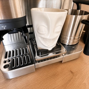 Könnte beinhalten: Ein weißer Keramikbecher mit einem skulptierten, ausdrucksstarken Gesicht mit großen Augen und einem breiten Lächeln, platziert auf der Abtropfschale einer Espressomaschine aus Edelstahl. Die Maschine umfasst ein Mahlwerk und ein Milchkännchen.