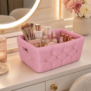 Op de afbeelding: Een rechthoekige roze opbergmand met een gecapitonneerde buitenkant met knoopdetails. De mand is gevuld met diverse cosmetische artikelen, waaronder verschillende make-upkwasten met bruine haren, huidverzorgingsflesjes en kleine potjes.