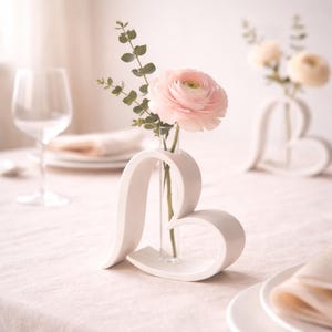 Könnte beinhalten: Eine weiße herzförmige Vase mit einer rosa Blume und Grünzeug. Die Vase steht auf einer hellrosa Tischdecke, mit einem Weinglas und Tellern im Hintergrund. Die Vase ist ein dekorativer Gegenstand.