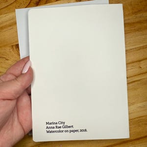 Puede incluir: Una tarjeta en blanco, blanquecina, con esquinas redondeadas, sostenida sobre un sobre gris. La tarjeta presenta el texto "Marina City, Anna Rae Gilbert, Acuarela sobre papel, 2018."