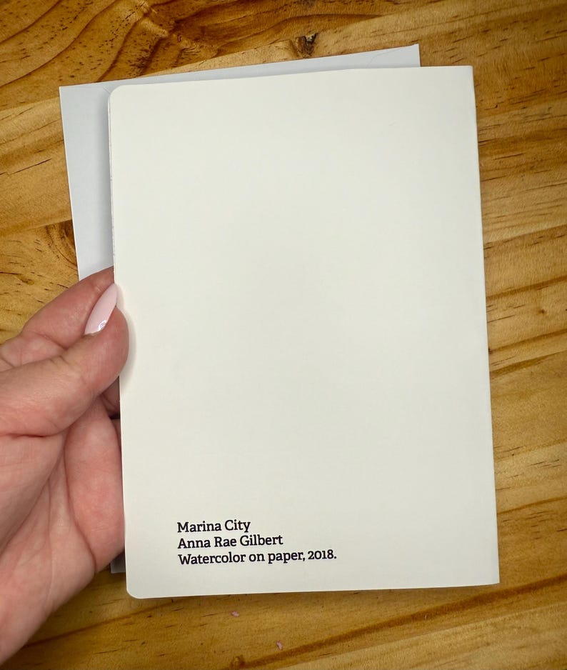 Puede incluir: Una tarjeta en blanco, blanquecina, con esquinas redondeadas, sostenida sobre un sobre gris. La tarjeta presenta el texto "Marina City, Anna Rae Gilbert, Acuarela sobre papel, 2018."