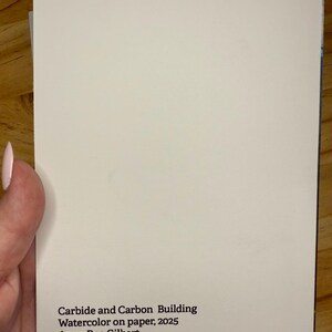 Puede incluir: Un papel rectangular blanquecino con el texto "Carbide and Carbon Building, Watercolor on paper, 2025, Anna Rae Gilbert" impreso en negro. El papel est&aacute; sostenido por una persona.