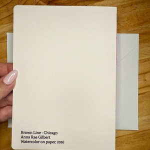 Puede incluir: Una tarjeta color crema con el texto "Brown Line - Chicago Anna Rae Gilbert Watercolor on paper, 2016" se sostiene junto a un sobre gris claro. La tarjeta y el sobre est&aacute;n sobre una superficie de madera.