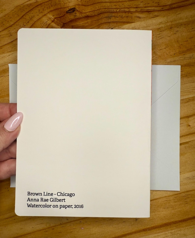 Puede incluir: Una tarjeta color crema con el texto "Brown Line - Chicago Anna Rae Gilbert Watercolor on paper, 2016" se sostiene junto a un sobre gris claro. La tarjeta y el sobre est&aacute;n sobre una superficie de madera.