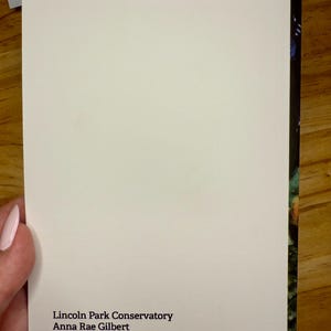 Puede incluir: Una tarjeta rectangular blanca con esquinas redondeadas, sostenida por una mano. La tarjeta presenta el texto: "Lincoln Park Conservatory, Anna Rae Gilbert, Watercolor on paper, 2016." El borde derecho muestra un vistazo de una imagen colorida.