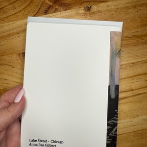Puede incluir: Una tarjeta de felicitaci&oacute;n blanca con el texto "Lake Street - Chicago Anna Rae Gilbert Oil on canvas, 2015" se sostiene contra una superficie de madera. La tarjeta tiene una peque&ntilde;a secci&oacute;n de una pintura en el lado derecho.