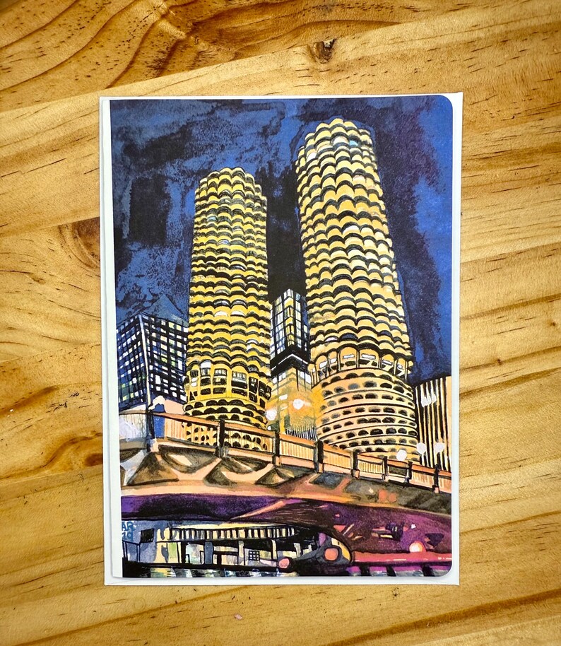 Puede incluir: Una tarjeta de felicitaci&oacute;n con una ilustraci&oacute;n de dos edificios altos e iluminados con un dise&ntilde;o circular &uacute;nico. Los edificios se alzan sobre un cielo nocturno azul oscuro, con un puente y otras estructuras urbanas en primer plano. La tarjeta est&aacute; sobre una superficie de madera.
