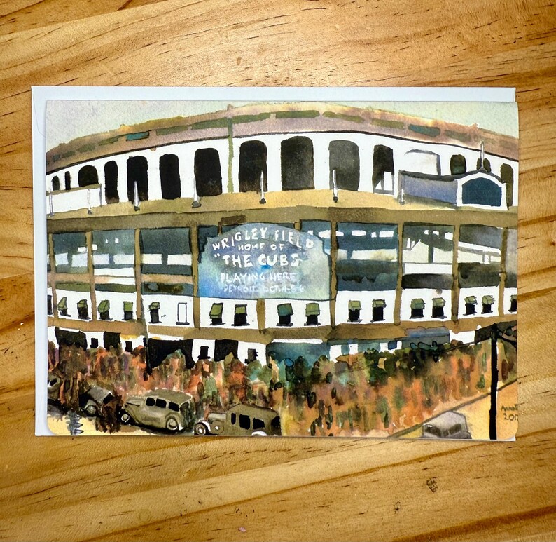 Puede incluir: Pintura a la acuarela del Wrigley Field, sede de los Cachorros de Chicago. La obra representa el exterior del estadio con el texto "Wrigley Field Home of the Cubs Playing Here" en un letrero. Coches antiguos aparcados delante.