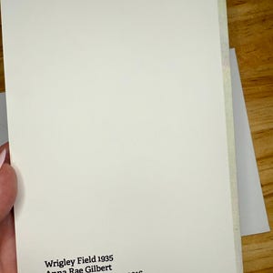 Puede incluir: Un cuaderno color crema con el texto "Wrigley Field 1935 Anna Rae Gilbert Watercolor on paper, 2016." El cuaderno est&aacute; sostenido en una mano y se encuentra sobre una superficie de madera.