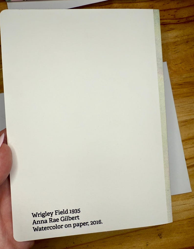 Puede incluir: Un cuaderno color crema con el texto "Wrigley Field 1935 Anna Rae Gilbert Watercolor on paper, 2016." El cuaderno est&aacute; sostenido en una mano y se encuentra sobre una superficie de madera.