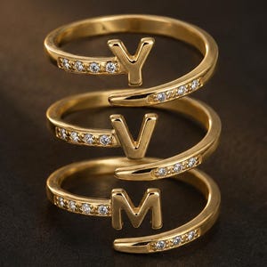 May include: A gold-toned spiral ring featuring three distinct tiers, each adorned with a single letter Y, V, and M respectively. The band is embellished with small, clear, sparkling stones set along the outer edges of the spiral design.