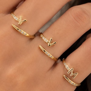 May include: Three gold-toned open rings, each featuring a single letter Y, V, or M. The bands are partially encrusted with small, clear, sparkling stones that extend from the letter towards the tapered end of the ring, creating a delicate look.
