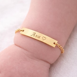 Pode incluir: Uma pulseira de identificação em metal dourado com uma placa retangular gravada com o nome Ava seguido por um pequeno símbolo de coração. A placa está presa a uma delicada corrente de elos dourada que envolve um pulso.