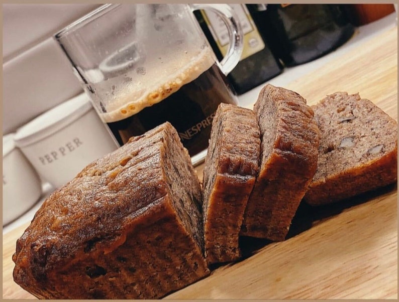 May include: A close-up of a wooden cutting board with slices of banana bread. The bread is light brown and has a slightly crumbly texture. There is a glass of coffee in the background.