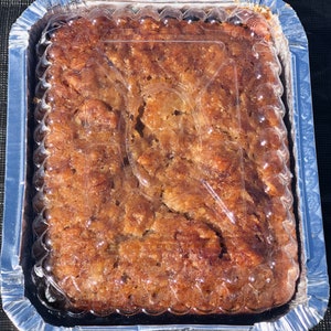 May include: A close-up of a rectangular pan of banana bread with a clear plastic lid. The bread is a light brown color and has a slightly crispy top.