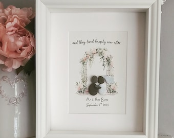 May include: A white framed print with a watercolor illustration of a floral archway. Two figures made of gray and brown stones are standing under the archway. The text "...and they lived happily ever after" is above the archway. The text "Mr & Mrs Barnes September 2nd 2023" is below the archway.