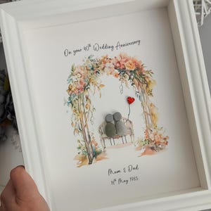 May include: A white framed picture with a watercolour illustration of a floral archway with two pebble people sitting on a bench. The text reads "On your 40th Wedding Anniversary", "Mum & Dad", and "18th May 1985".