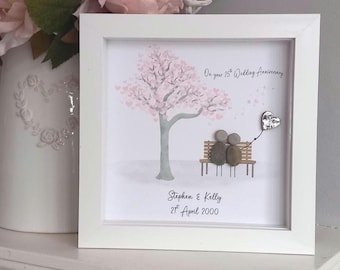 May include: A square white framed wall art piece featuring a printed illustration of a tree with pink leaves and a bench. Two pebble figures sit on the bench holding a silver heart balloon. Text reads: On your 25th Wedding Anniversary, Stephen & Kelly, 21st April 2000.