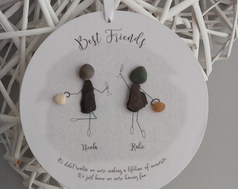 May include: White round plaque with the words "Best Friends" written in brown cursive. Two stick figures made of stones are holding hands. The names "Nicola" and "Kalie" are written below the figures. The quote "We didn't realize we were making a lifetime of memories. We just knew we were having fun." is written below the names.