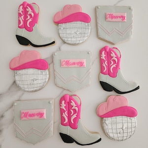 May include: Assortment of decorated sugar cookies in a Western theme. Designs include pink cowboy boots, disco ball shapes with pink cowboy hats, and gray pocket shapes with the name "Maevery" in pink.