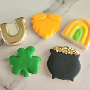 May include: Five decorated sugar cookies with a St. Patrick's Day theme. The cookies are shaped like a horseshoe, a shamrock, a pot of gold, a rainbow, and a leprechaun's beard.
