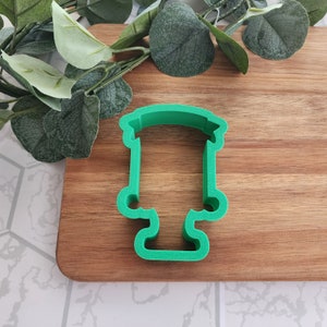May include: A bright green cookie cutter in the shape of a trophy. The cutter has a rectangular top, a long, straight body, and a curved base with two small handles. The cutter is made of plastic and is sitting on a wooden cutting board.