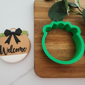 Puede incluir: Un cortador de galletas de pl&aacute;stico verde con forma de adorno navide&ntilde;o con un lazo y follaje. El cortador de galletas est&aacute; sobre una superficie de madera.