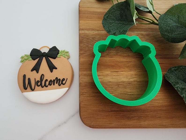 Puede incluir: Un cortador de galletas de pl&aacute;stico verde con forma de adorno navide&ntilde;o con un lazo y follaje. El cortador de galletas est&aacute; sobre una superficie de madera.
