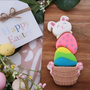 May include: A white bunny cookie with pink ears and a pink nose, holding a stack of three decorated Easter egg cookies. The bottom egg cookie is in a brown basket cookie. The cookies are on a wooden surface with a sign that says "Happy Easter".