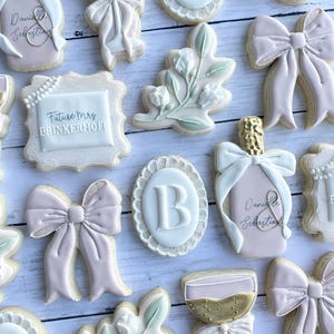 May include: A set of 12 decorated sugar cookies featuring a variety of designs, including a champagne flute, a bottle of champagne, a floral sprig, a bow, and the letter "B". The cookies are decorated with pink, white, and gold icing.