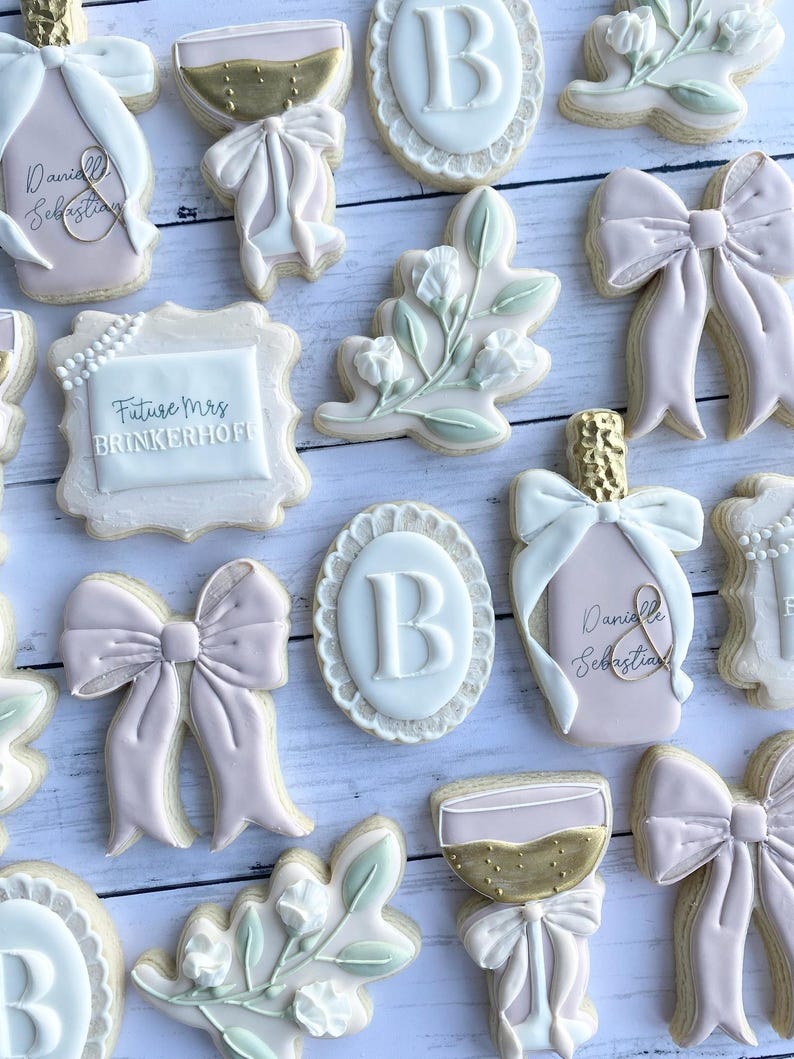 May include: A set of 12 decorated sugar cookies featuring a variety of designs, including a champagne flute, a bottle of champagne, a floral sprig, a bow, and the letter "B". The cookies are decorated with pink, white, and gold icing.