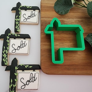 Puede incluir: Tres galletas decoradas con glaseado negro y verde, con forma de casa con un cartel que dice "Sold". Un cortador de galletas de pl&aacute;stico verde con forma de casa con un cartel est&aacute; sobre una tabla de cortar de madera.
